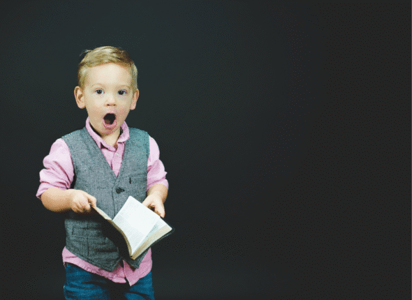 Aha Moments: young boy in pink shirt, gray vest, and jeans holding a book and with mouth open in surprise