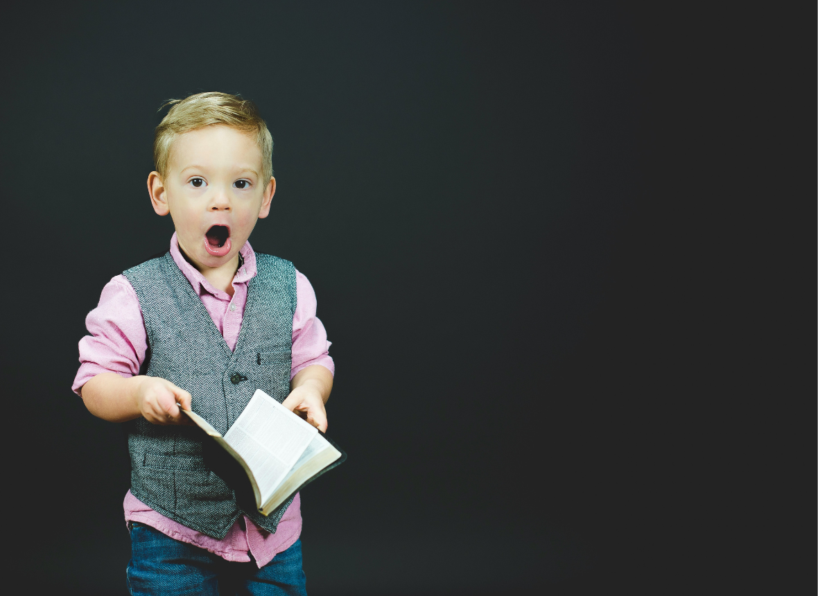 Aha Moments: young boy in pink shirt, gray vest, and jeans holding a book and with mouth open in surprise