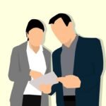 Initial Consultation - image of a woman, with black hair and wearing a white shirt and a gray blazer, talking to a man, with black hair and wearing a gray shirt and a black blazer, both looking at a document