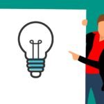 Workshops - In Person: Image of a woman wearing a red blouse, a black jacket, and blue pants pointing at a whiteboard showing a large lightbulb - signifying the sharing of the ideas through presentation